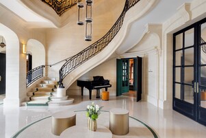 Un lobby majestuoso con una escalera de mármol, un piano de cola y una mesa de centro de vidrio con un jarrón de flores.