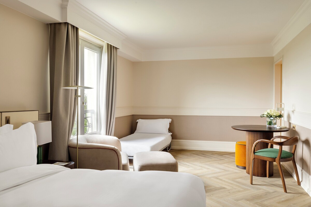 Habitación de hotel con cama, dos mesitas de noche, una silla, una mesita con un jarrón de flores y una ventana con cortinas.