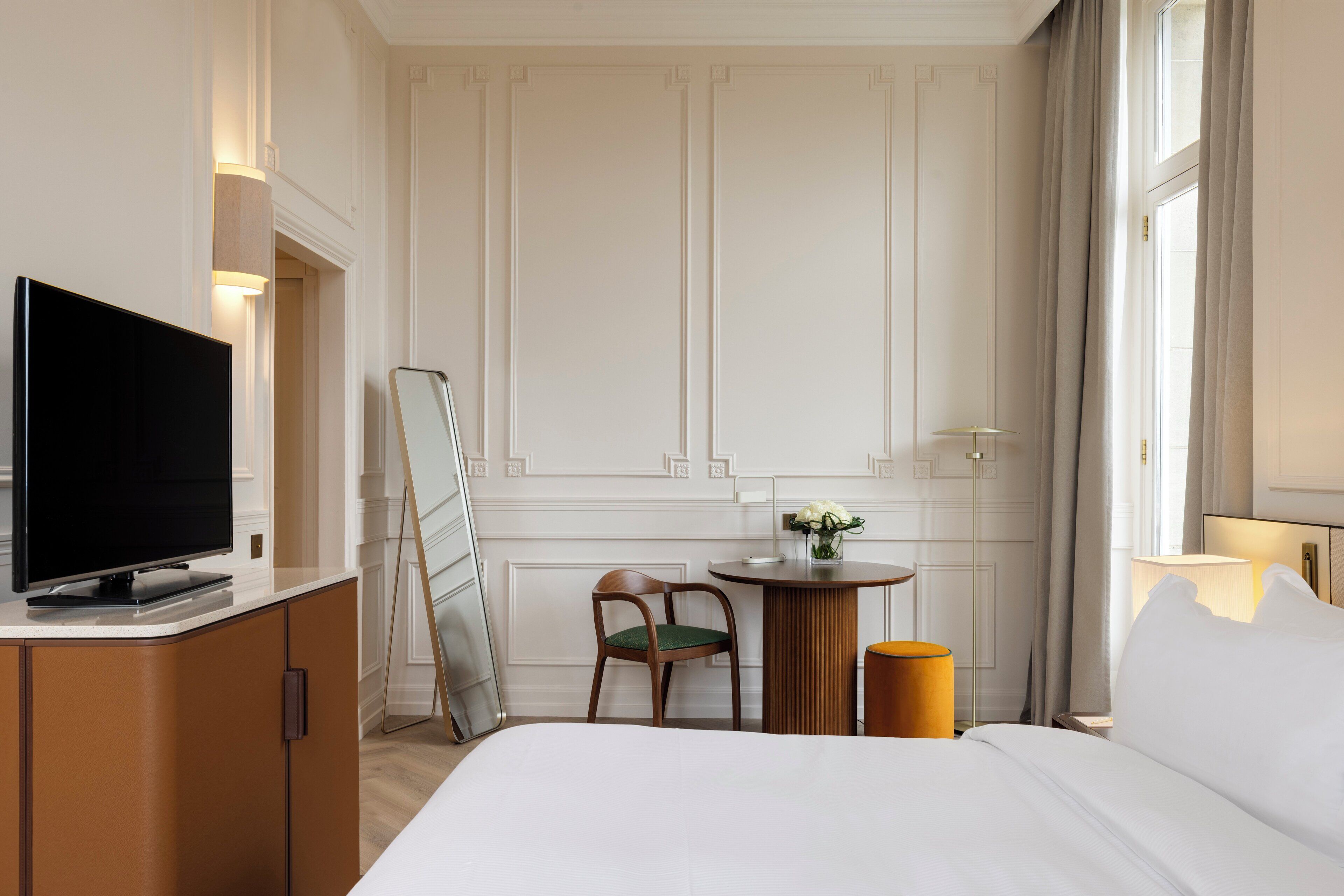A hotel room with a bed, a television on a cabinet, a small table with a chair, and a window with curtains.
