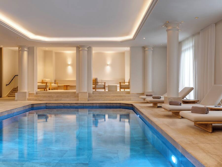 Una amplia piscina interior con superficie de agua azul cristalina, rodeada de sillones de descanso y una escalera que conduce a una zona de estar con mesas y sillas.