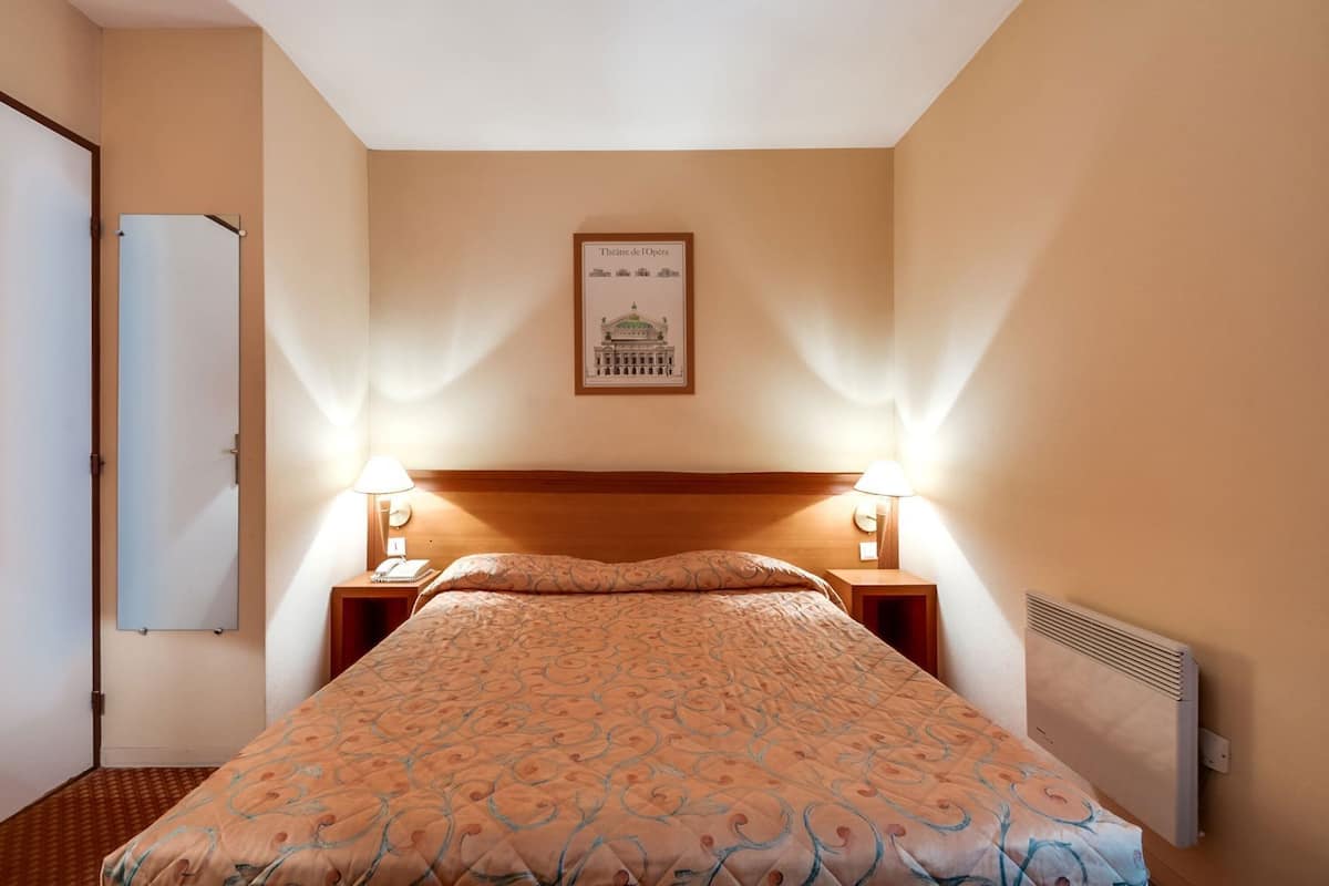 Habitación de hotel con cama, mesitas de noche, lámpara de pared, radiador y un cuadro con una vista de un edificio.
