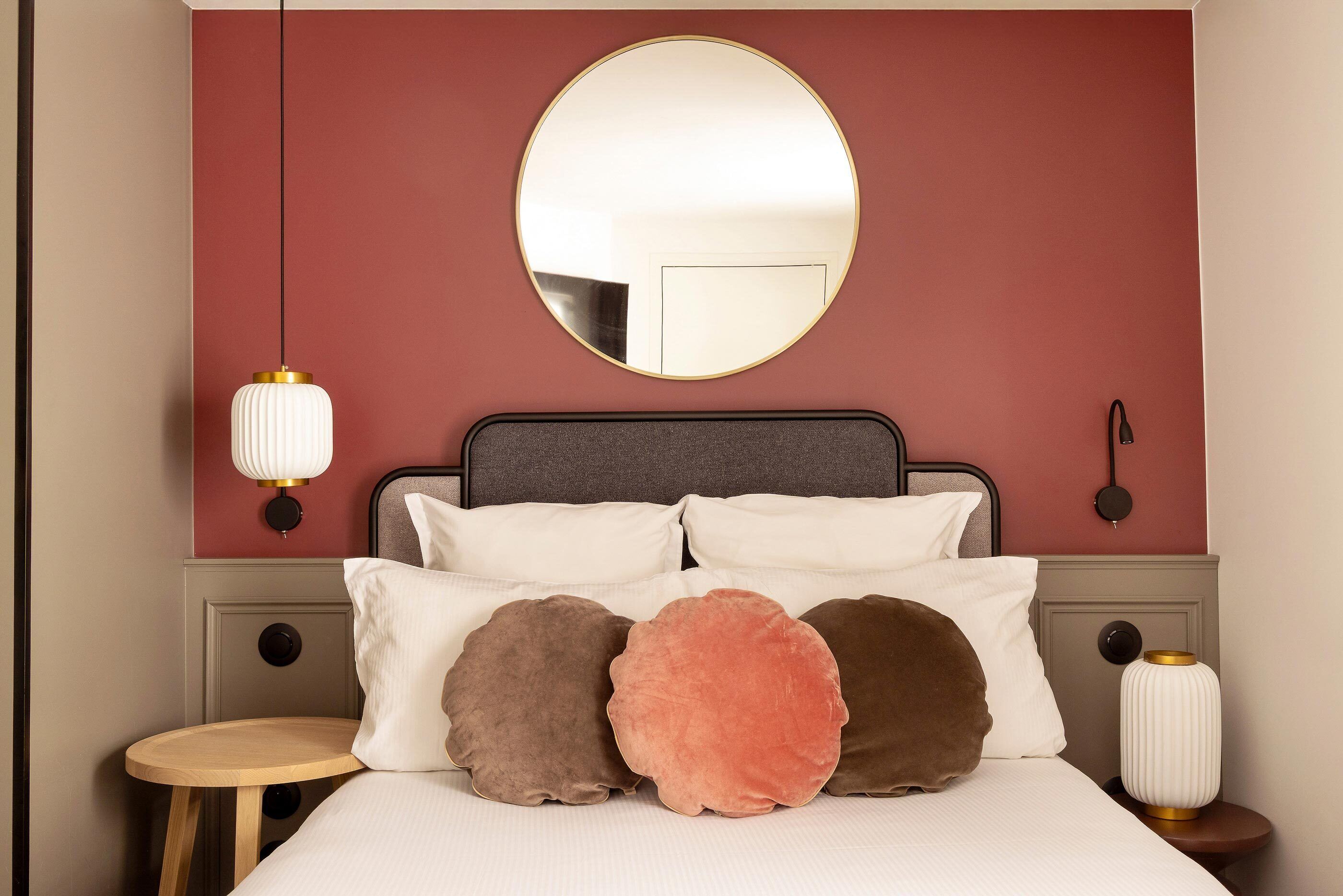 Une chambre avec un miroir rond, un lit avec de nombreux coussins, une table de chevet et une lampe fixée au mur.