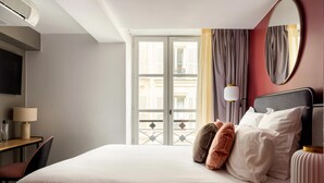 A hotel room with a large bed, a desk with a lamp, a TV, and a balcony with curtains. - La Dépendance (Paris)