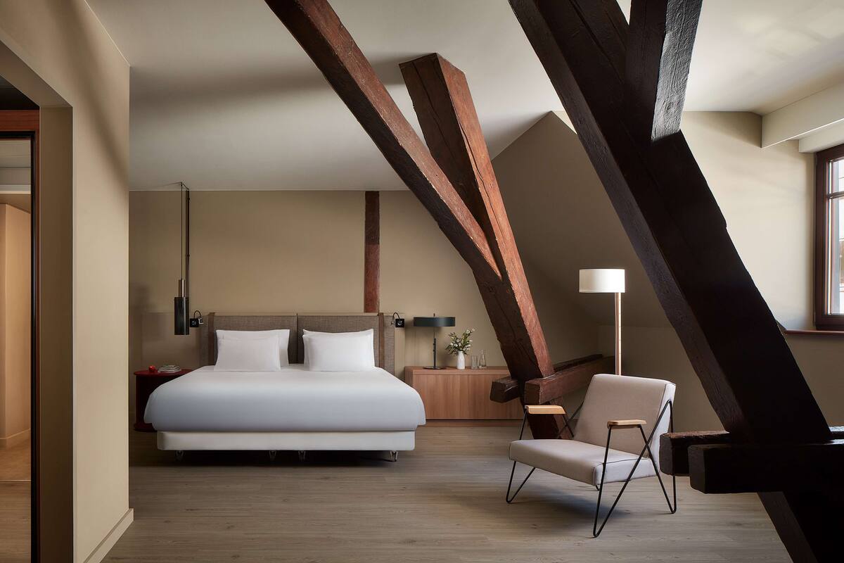 A modern hotel room with a large bed, a wooden chair, and exposed beams.