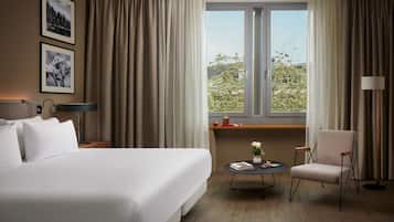 Una habitación de hotel con una cama grande, un escritorio con lámpara, una silla, una mesita con un florero y una ventana con cortinas.