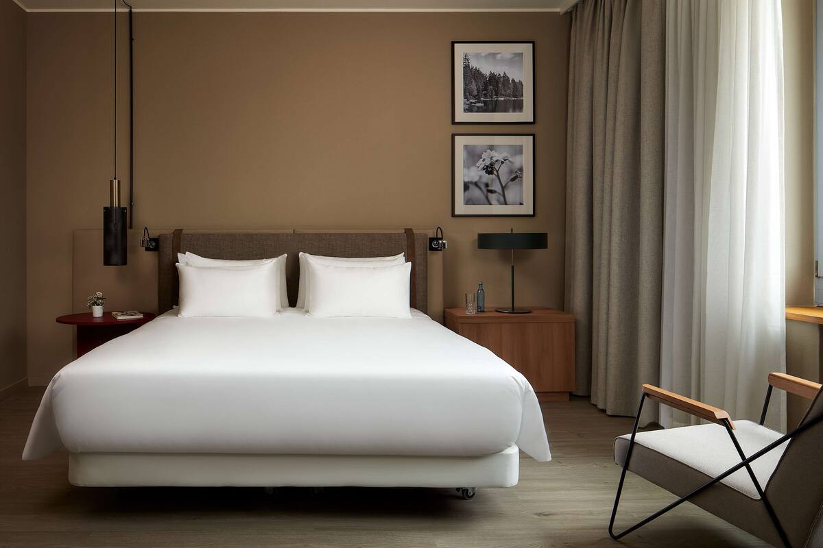 A modern hotel room with a large bed, two bedside tables, a chair, and two framed pictures on the wall.