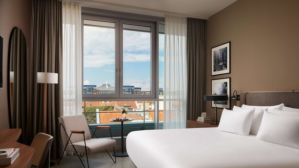 A hotel room with a large bed, a desk with a chair, a small table, and a view of the city through large windows.