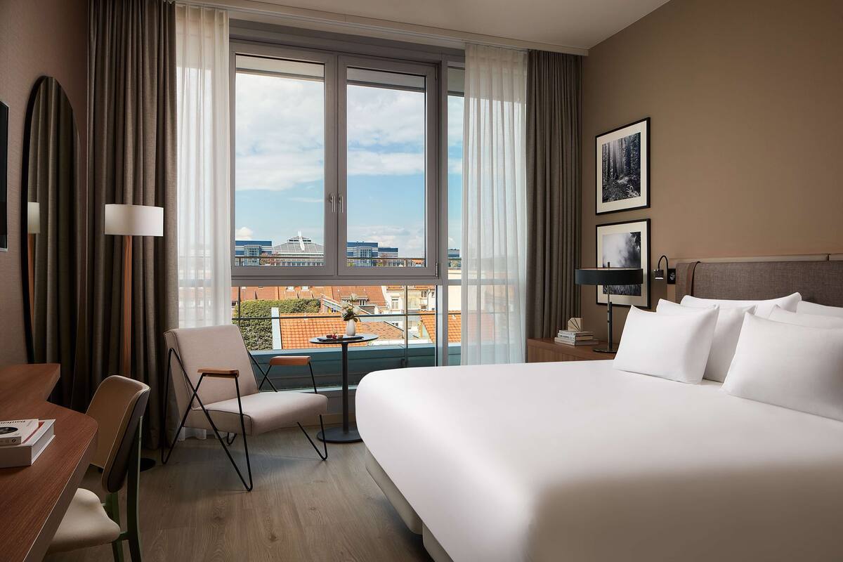 A hotel room with a large bed, a desk with a chair, a small table, and a view of the city through large windows.