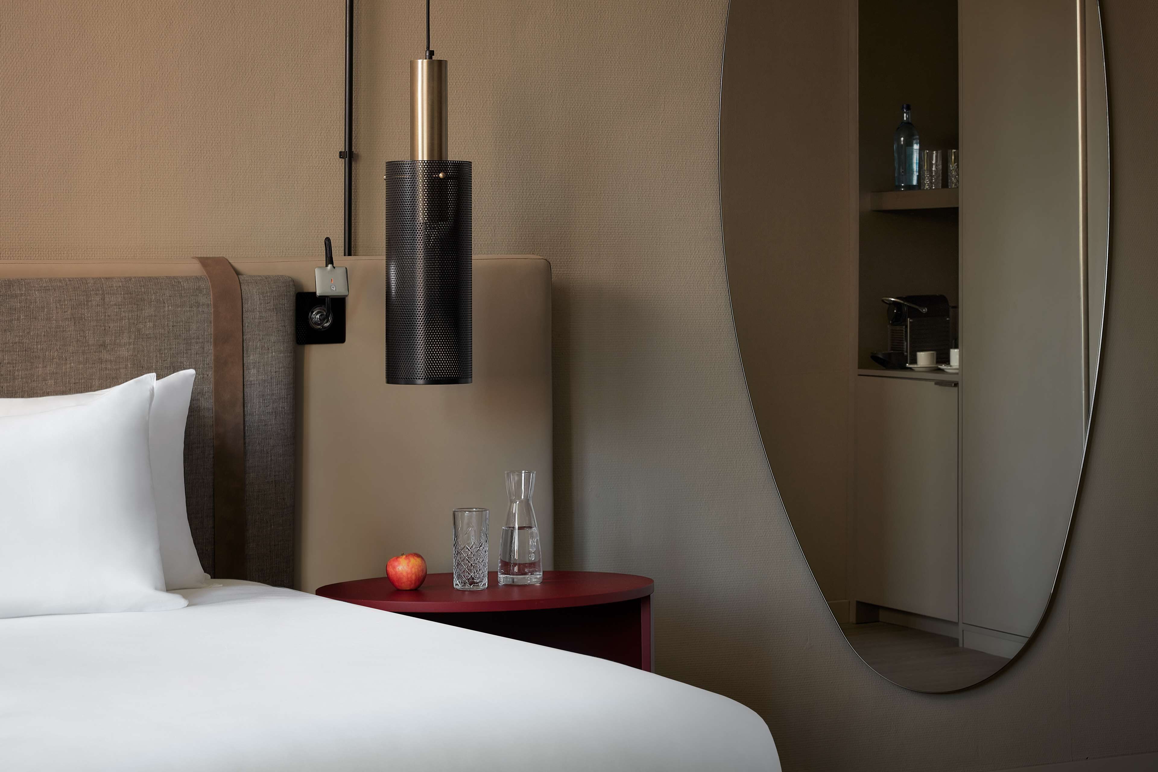 Una habitación de hotel con una cama, una mesa redonda con una mesita auxiliar roja, una lámpara colgante y un espejo.
