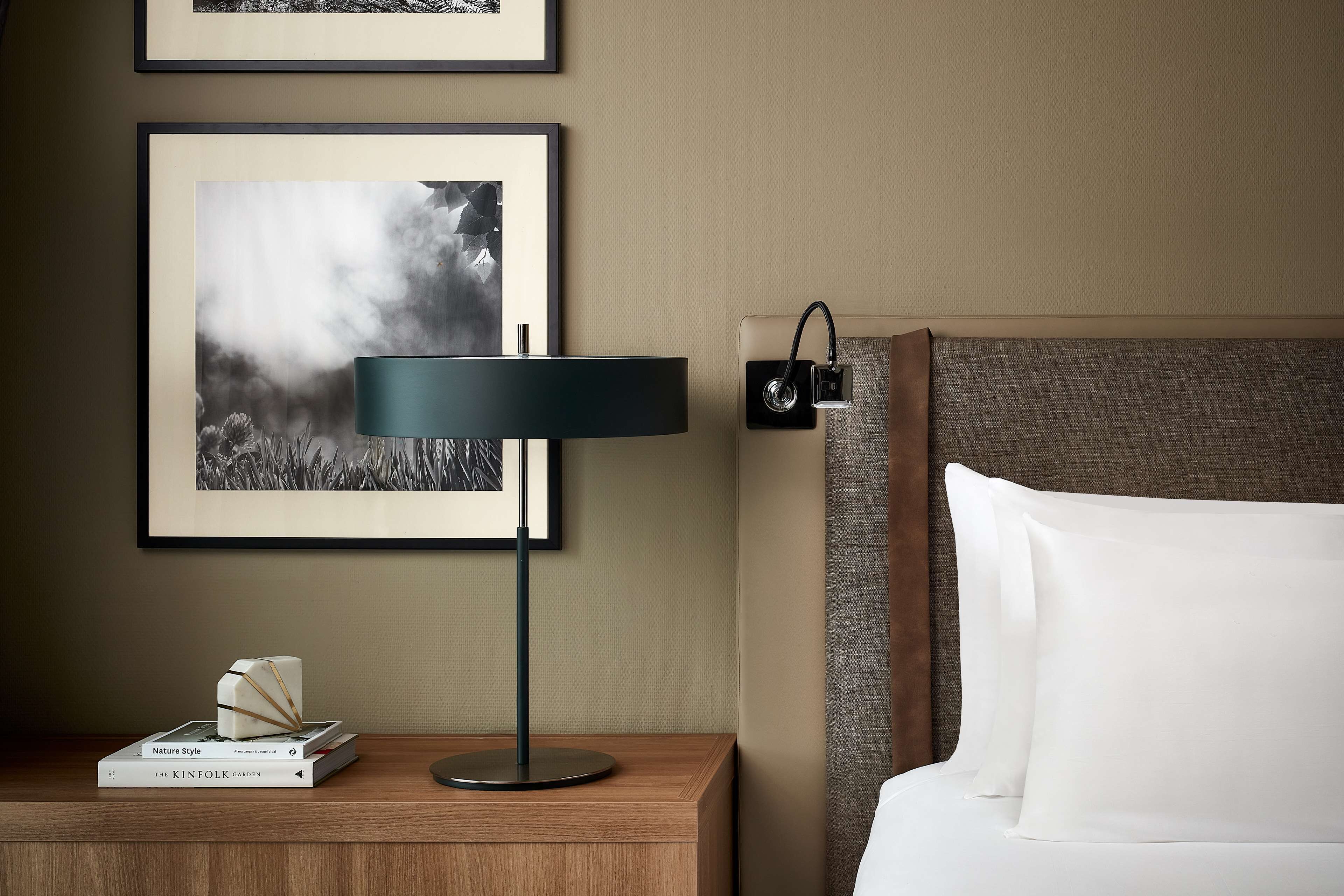 Habitación de hotel impecable con cama, mesita de noche con lámpara, cuadros en la pared y un libro titulado 