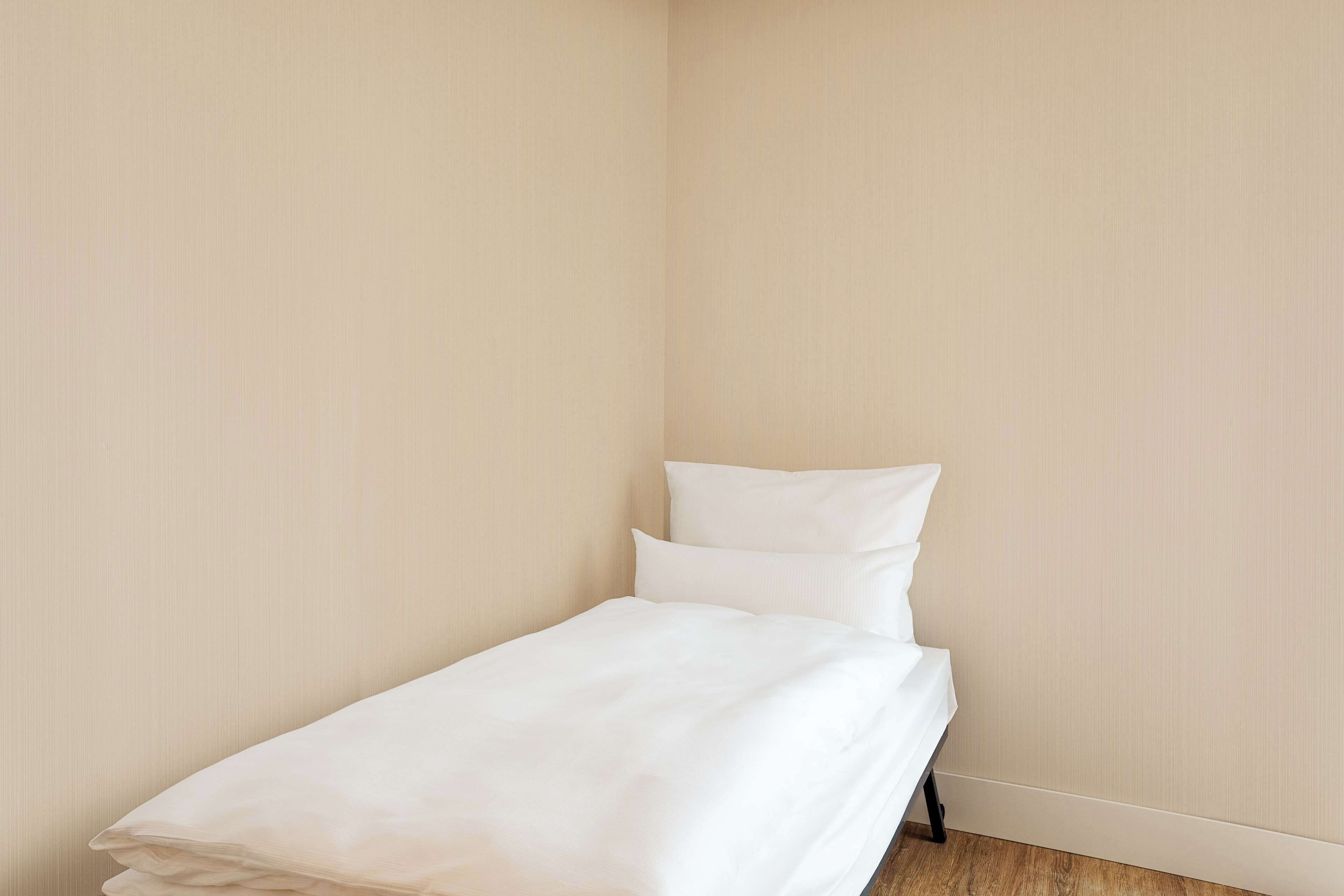 Ein kleines, ordentlich bezogenes Bett in einem Zimmer mit schlichten Wänden und Holzboden.