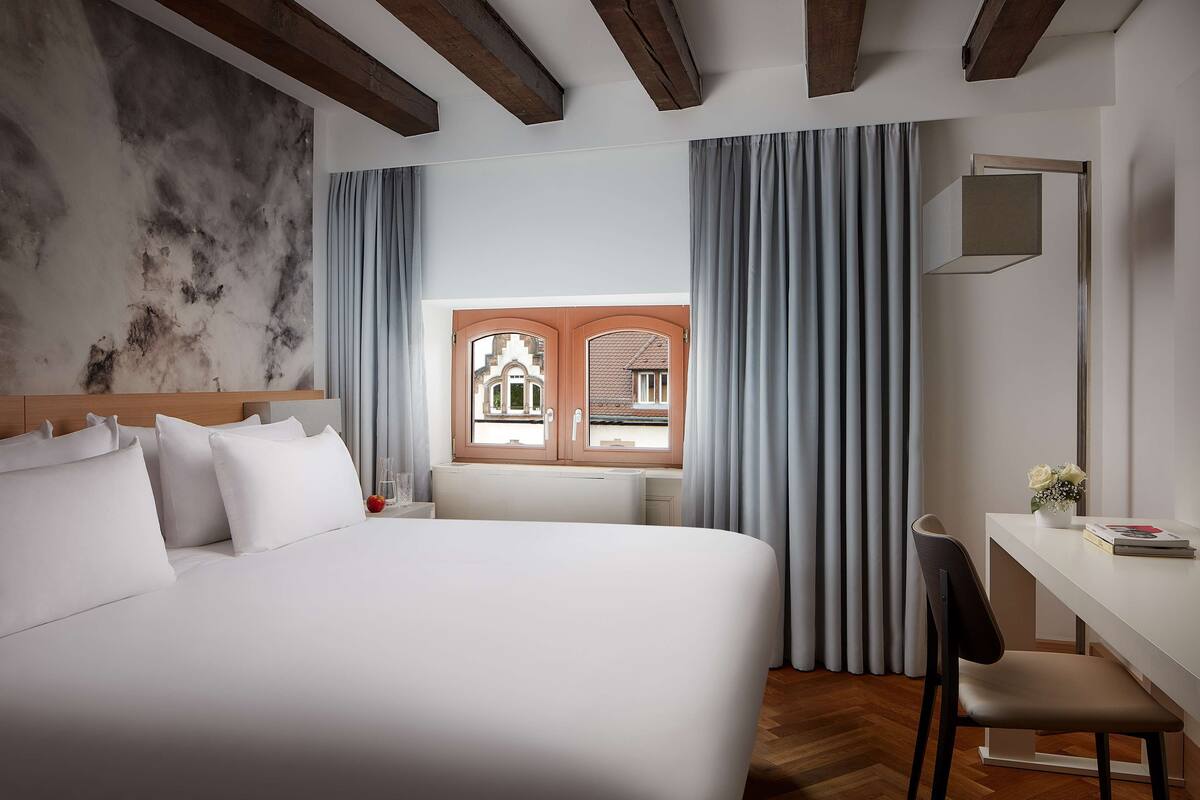 A hotel room with a large bed, a desk with a chair, a window with curtains, and a view of a building outside.