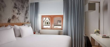 Habitación de hotel con una cama grande, un escritorio con silla, una ventana con cortinas y vistas a un edificio.