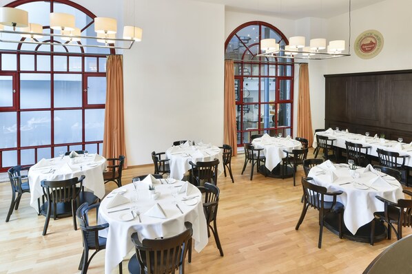 A dining area with multiple round tables set with white tablecloths and chairs, large windows with curtains, and a chandelier. - NH Collection Heidelberg (Heidelberg)