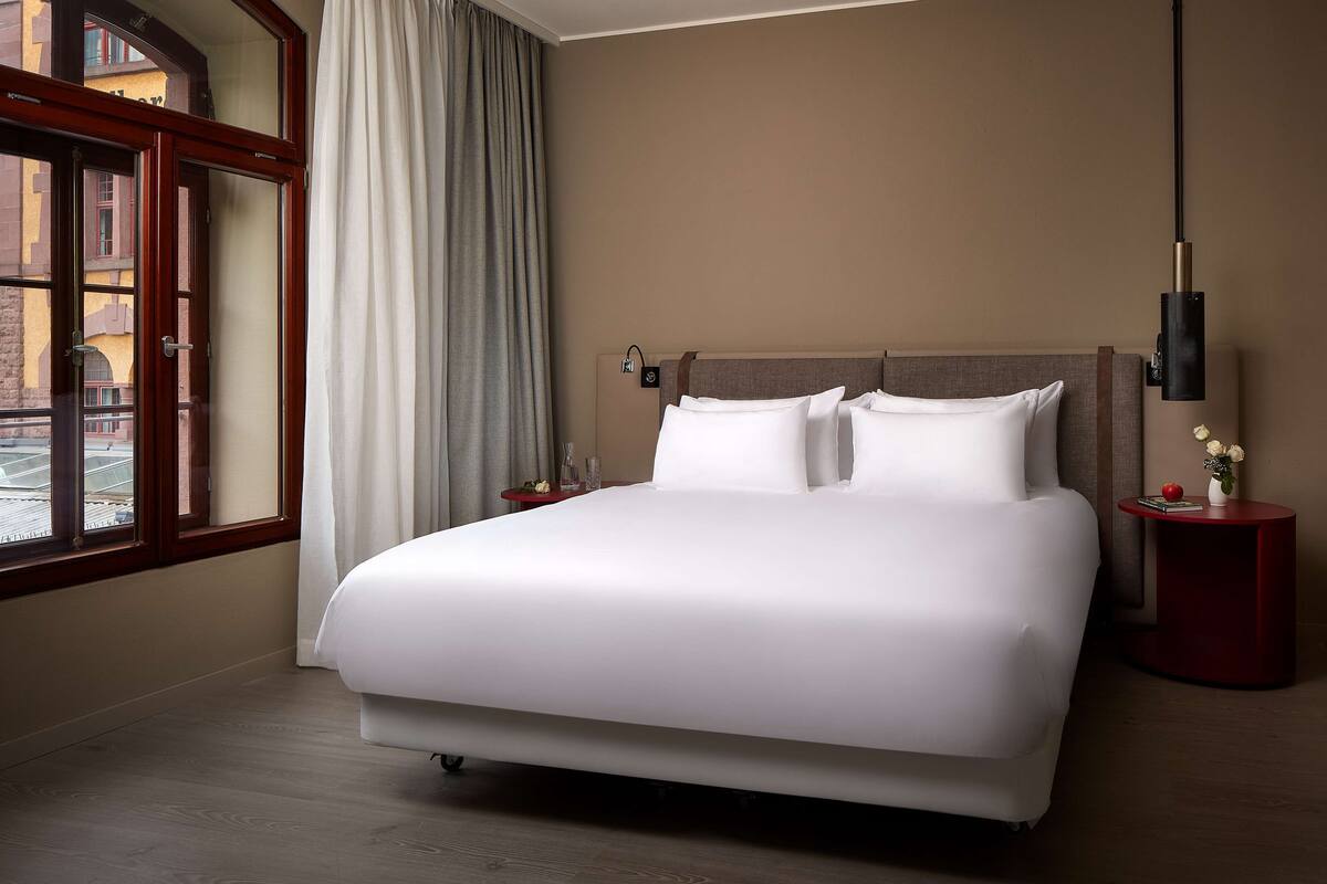 A hotel room with a large bed, a nightstand with a lamp, and a view of buildings through a window.