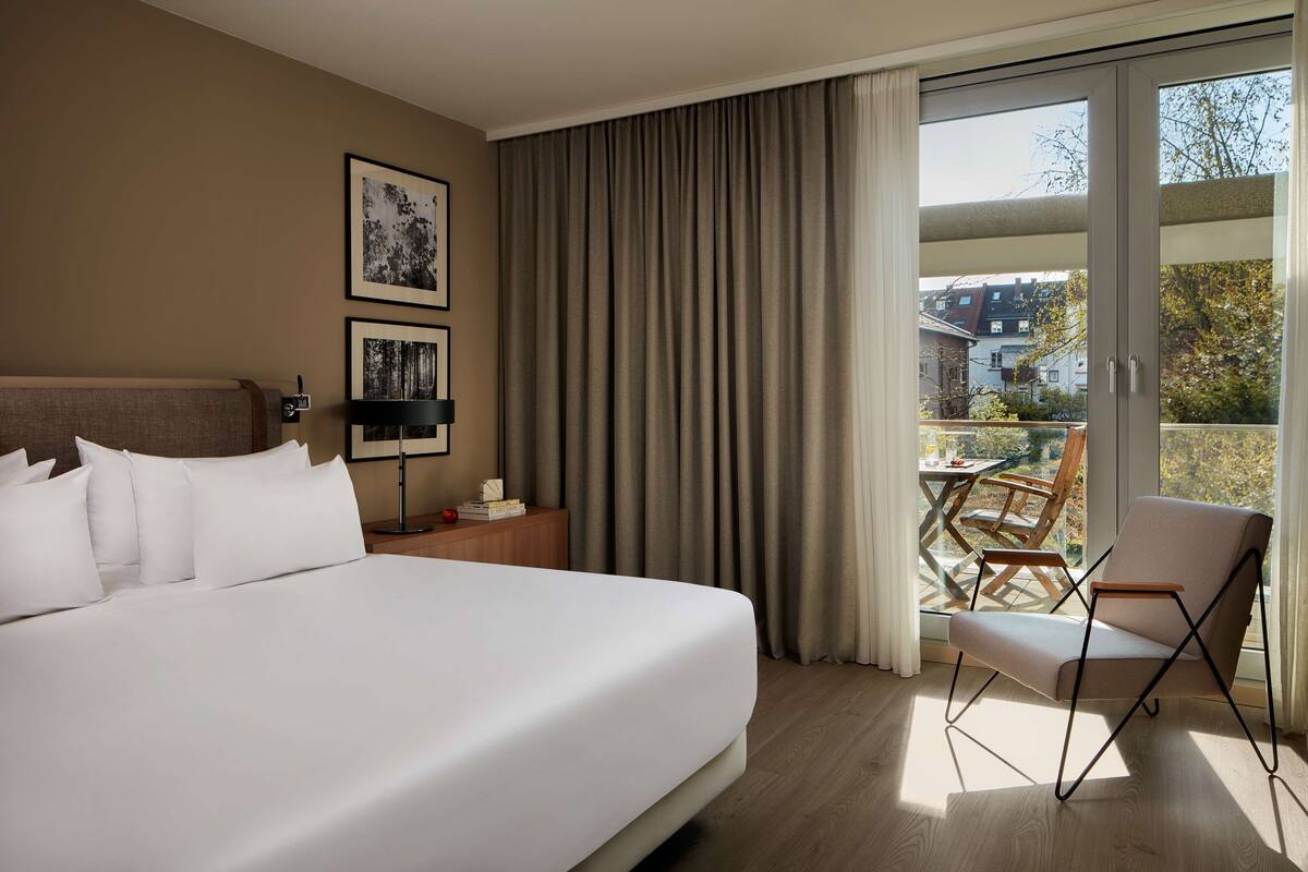A hotel room with a large bed, a chair, a table, and a balcony with a view of buildings and trees.