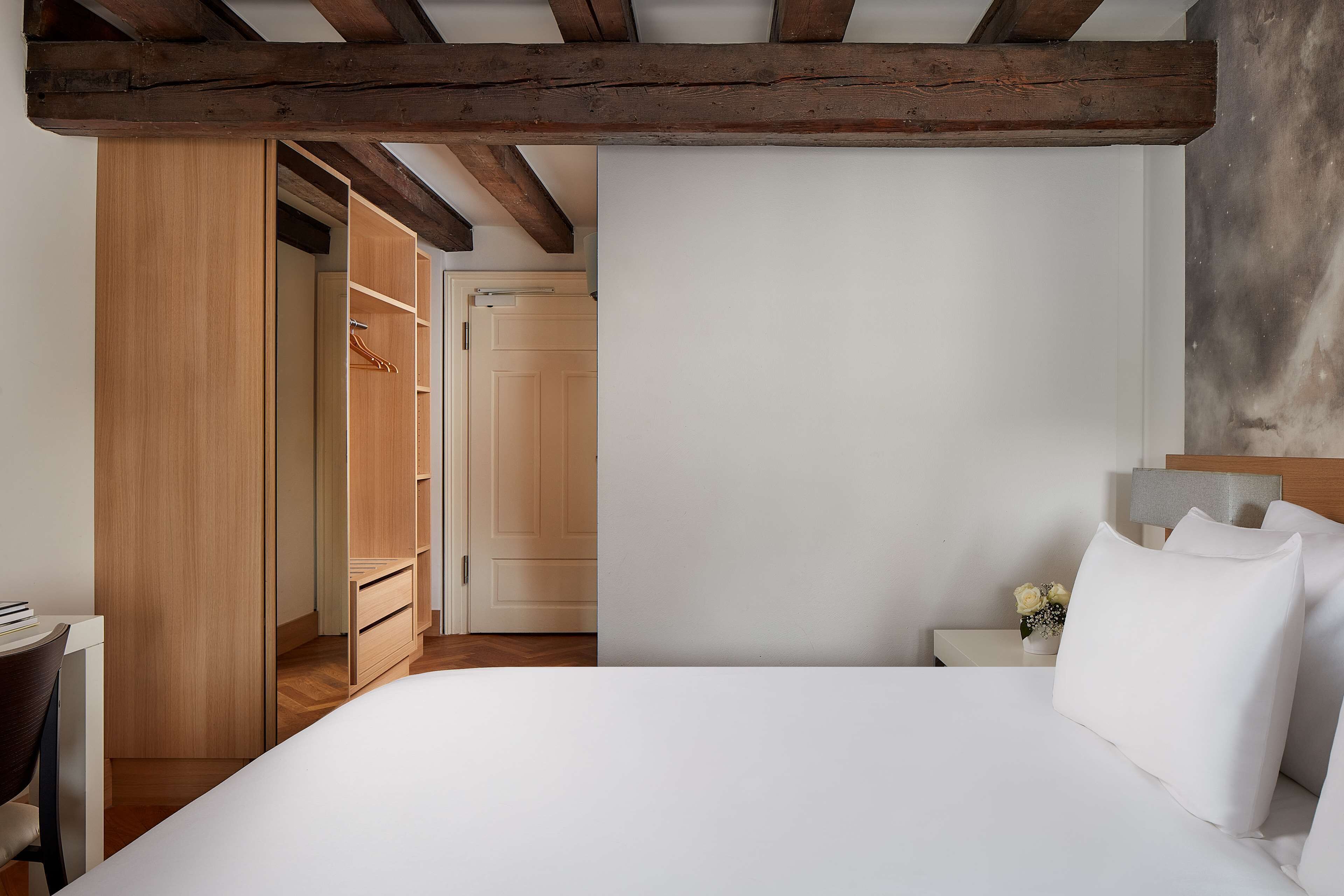 A bedroom with a wooden wardrobe, a bed with white linens, a nightstand with a lamp, and a small vase with flowers.
