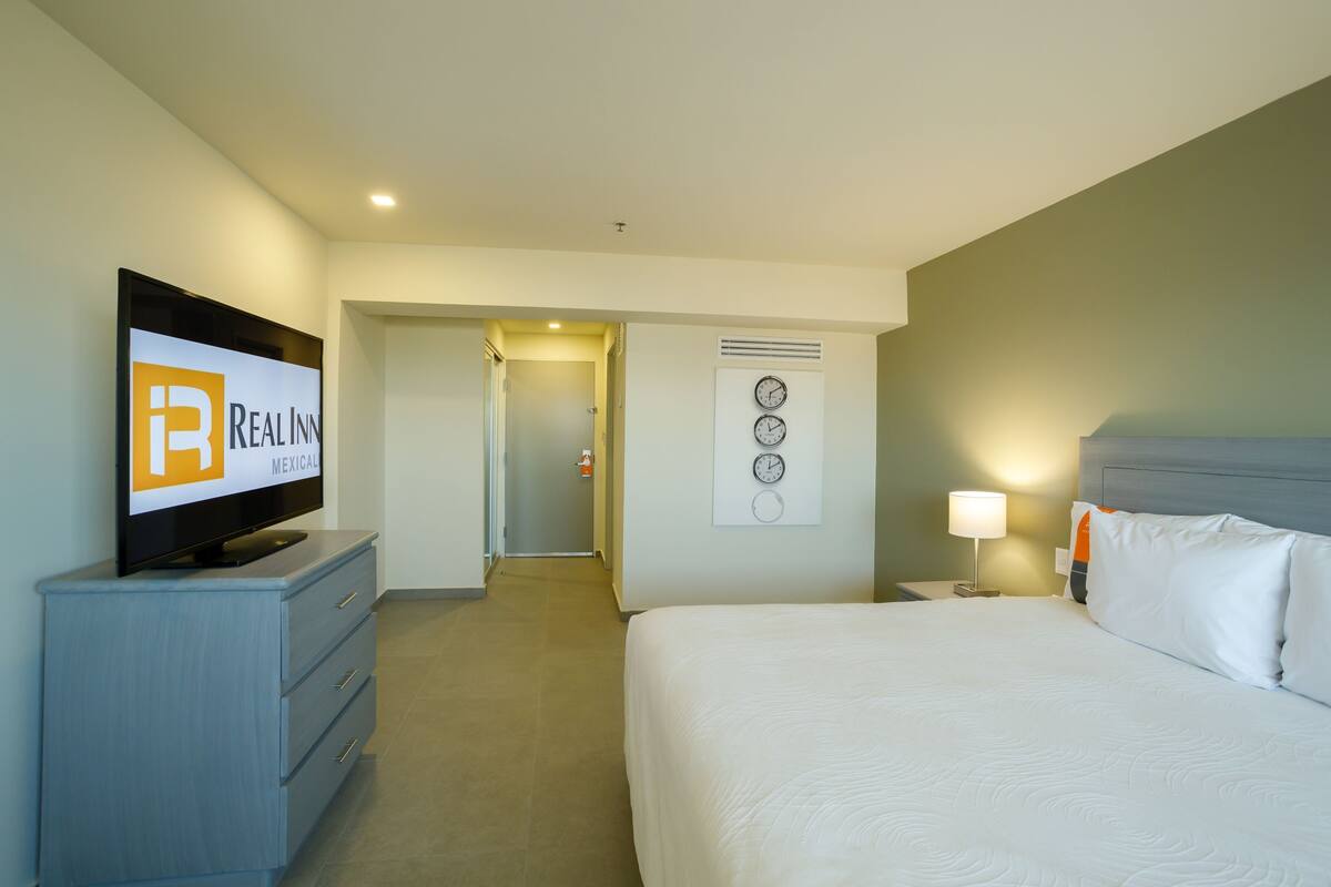 Habitación de hotel con una cama, un televisor en una cómoda, una mesita de noche con una lámpara y una puerta que da a otra habitación.