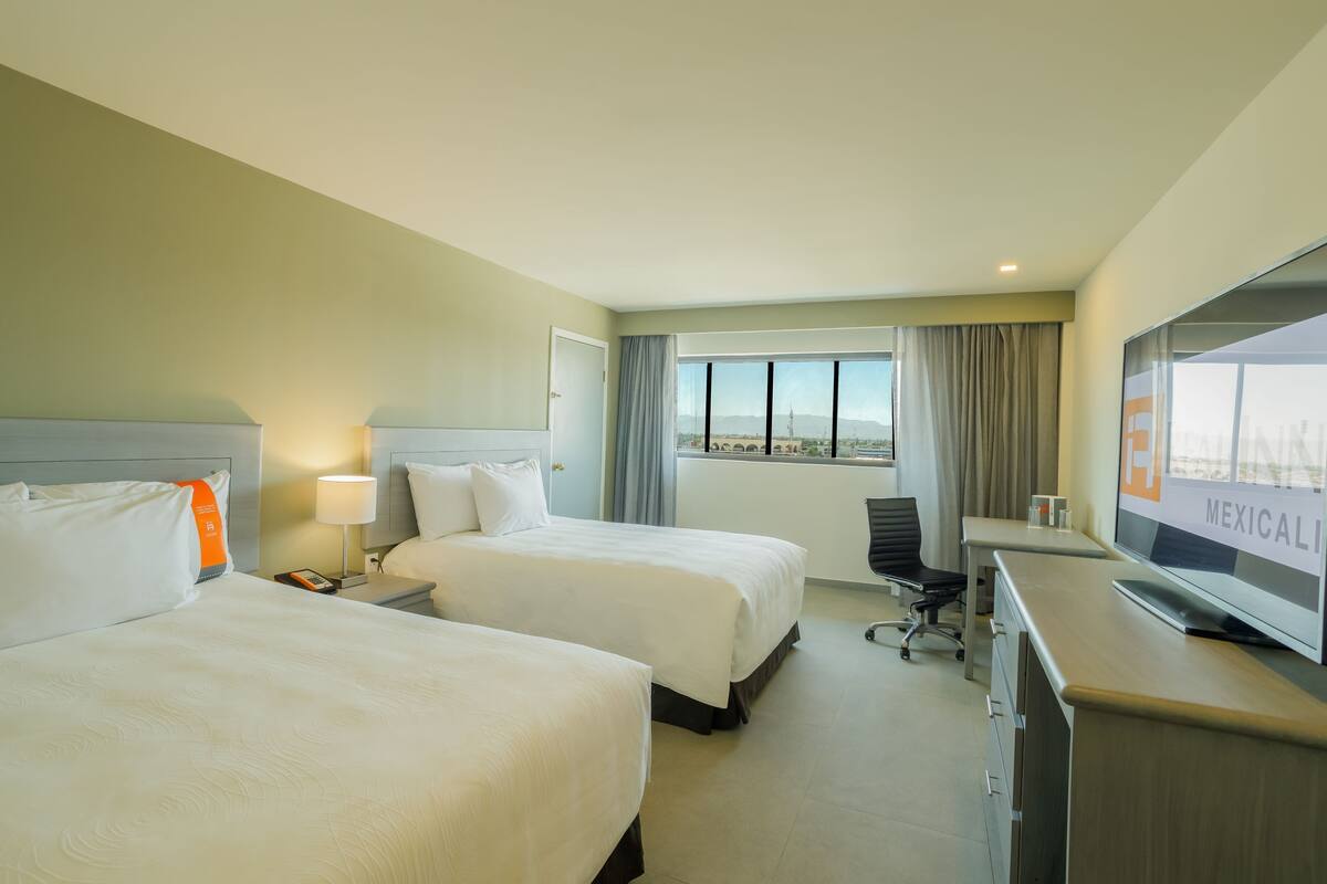 Habitación de hotel con dos camas, un escritorio, una silla, un televisor y ventana con vista a la ciudad.