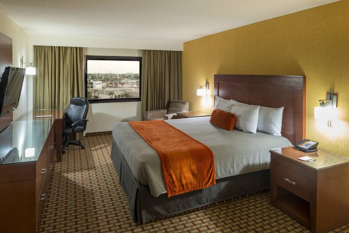 Habitación de hotel con cama, escritorio, silla, televisor y ventana con cortinas.