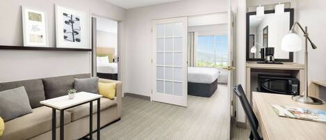 A hotel room with a sofa, a small table, a desk with a lamp, a microwave, and a large mirror. There is a door leading to a balcony with a view.
