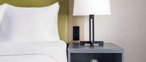 Un lit bien fait, avec du linge de lit blanc, une tête de lit verte, une table de chevet avec une lampe et une petite plante posée sur la table.