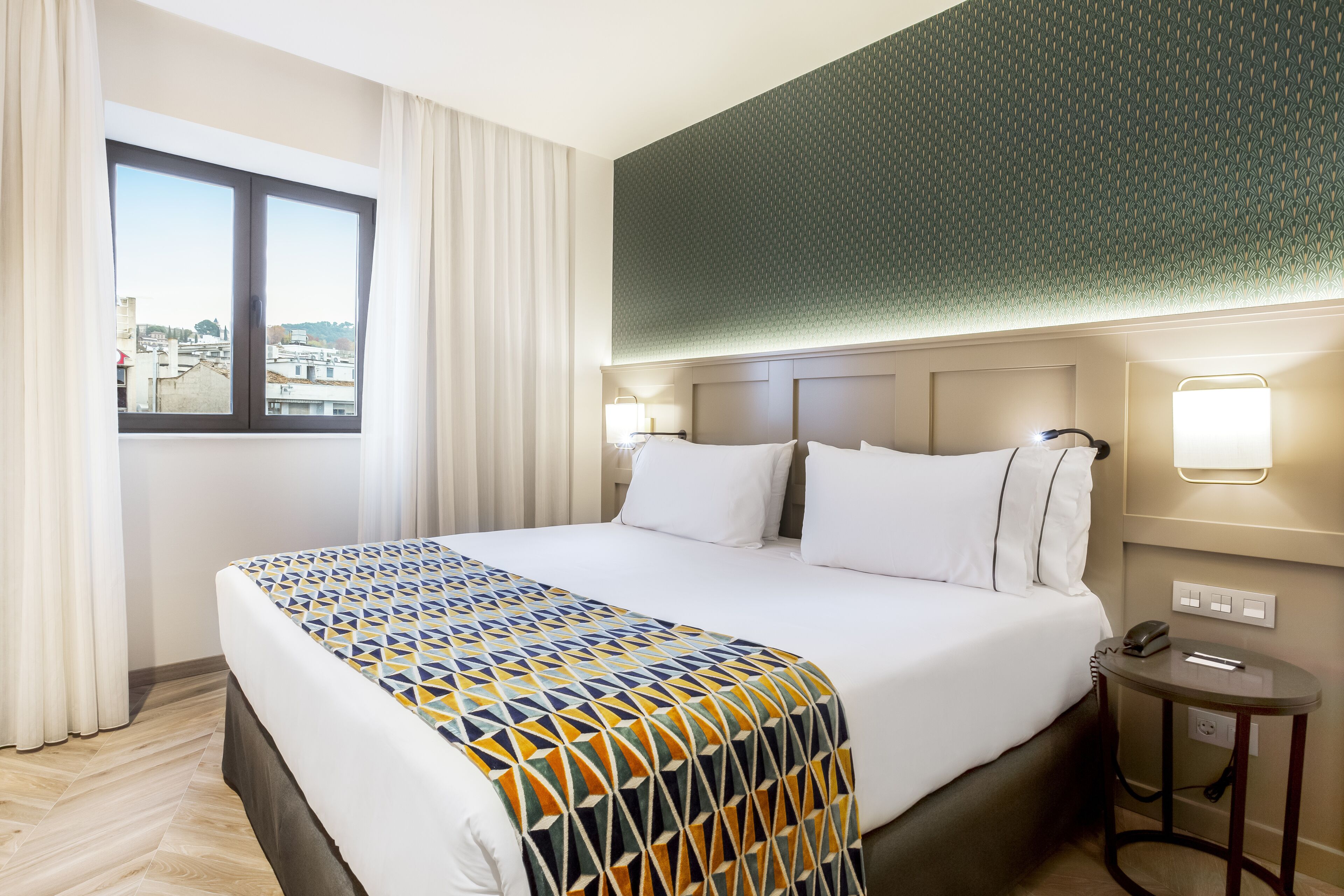 Une chambre d’hôtel avec un grand lit, une fenêtre avec des rideaux, un mur à motifs et une table de chevet avec une lampe.