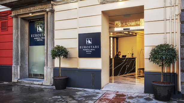 The entrance of the Eurostars Puerta Real Hotel with a glass door, potted plants, and a reception desk visible inside. - Eurostars Puerta Real Hotel (Granada)