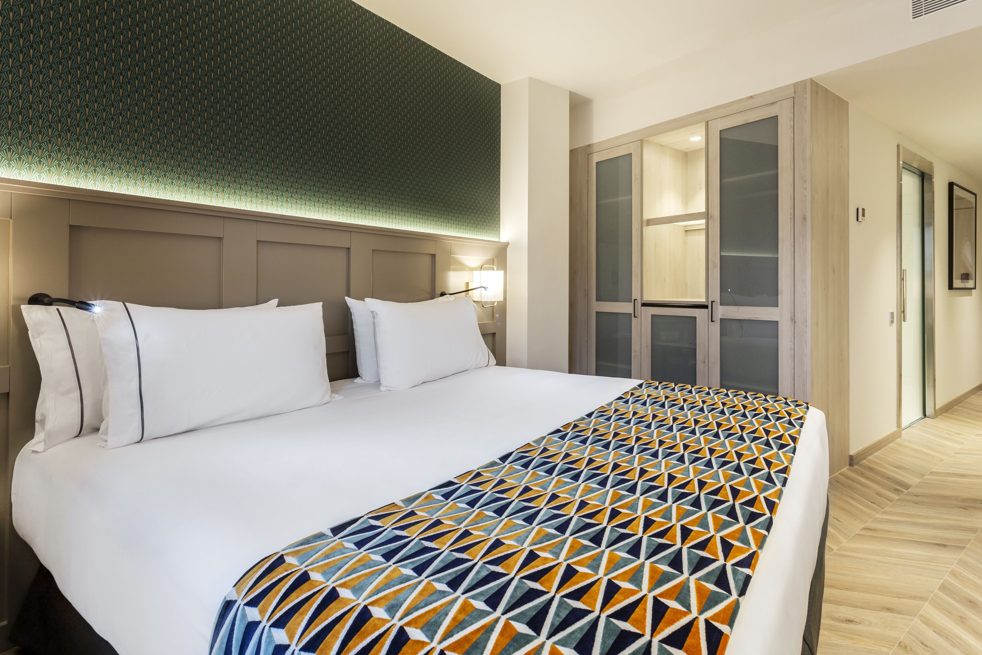 Una habitación de hotel con una cama grande, un plaid estampado geométricamente, un cabecero con iluminación integrada, un vestidor y suelo de madera.