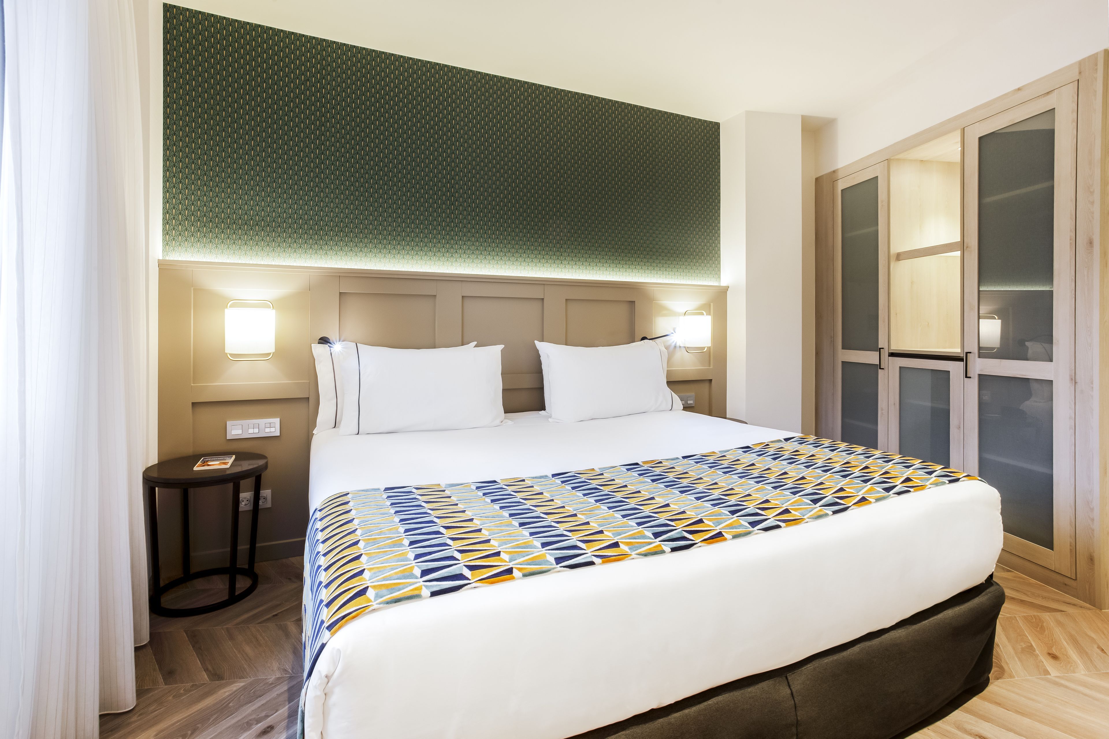 Quarto de hotel com cama, abajures, uma mesinha e um guarda-roupa com portas de vidro.
