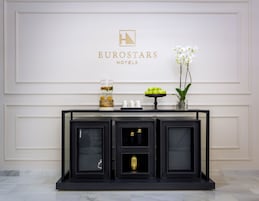 Un mostrador de recepción con el logotipo de Eurostars Hotels, una fuente de agua, una exposición de frutas y un jarrón con flores blancas.