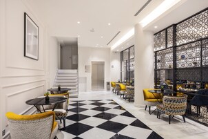 Un moderno vestíbulo de hotel con piso de cuadros, una escalera y un área de descanso con sillas y mesas amarillas.