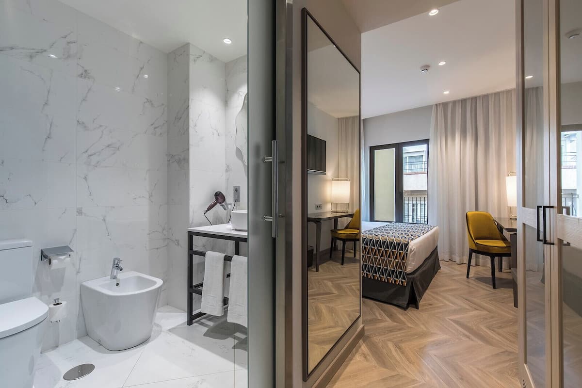 A modern hotel room with a marble bathroom, a bed with a patterned headboard, a desk with a chair, and a view through a glass door.