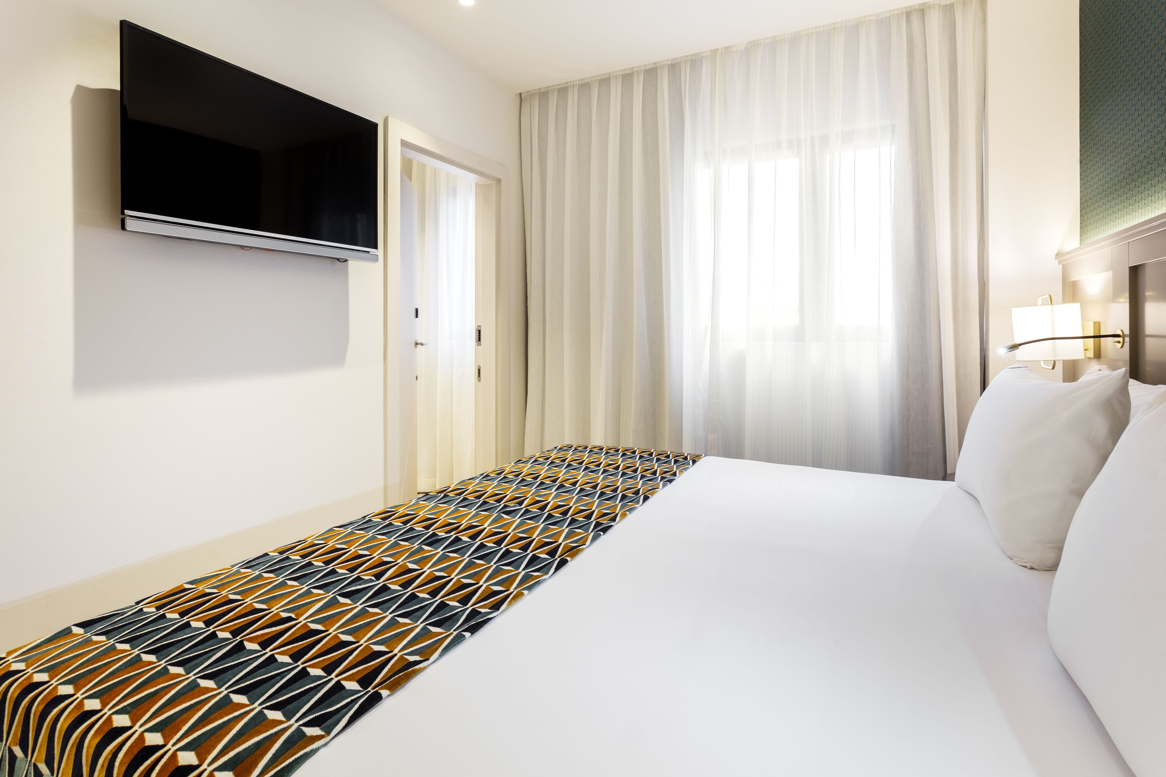 Una habitación de hotel con una cama grande, un televisor de pantalla plana montado en la pared, una ventana con cortinas traslúcidas y una lámpara de noche.