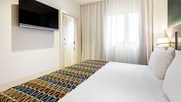 Una habitación de hotel con una cama grande, un televisor de pantalla plana montado en la pared, una ventana con cortinas traslúcidas y una lámpara de noche.