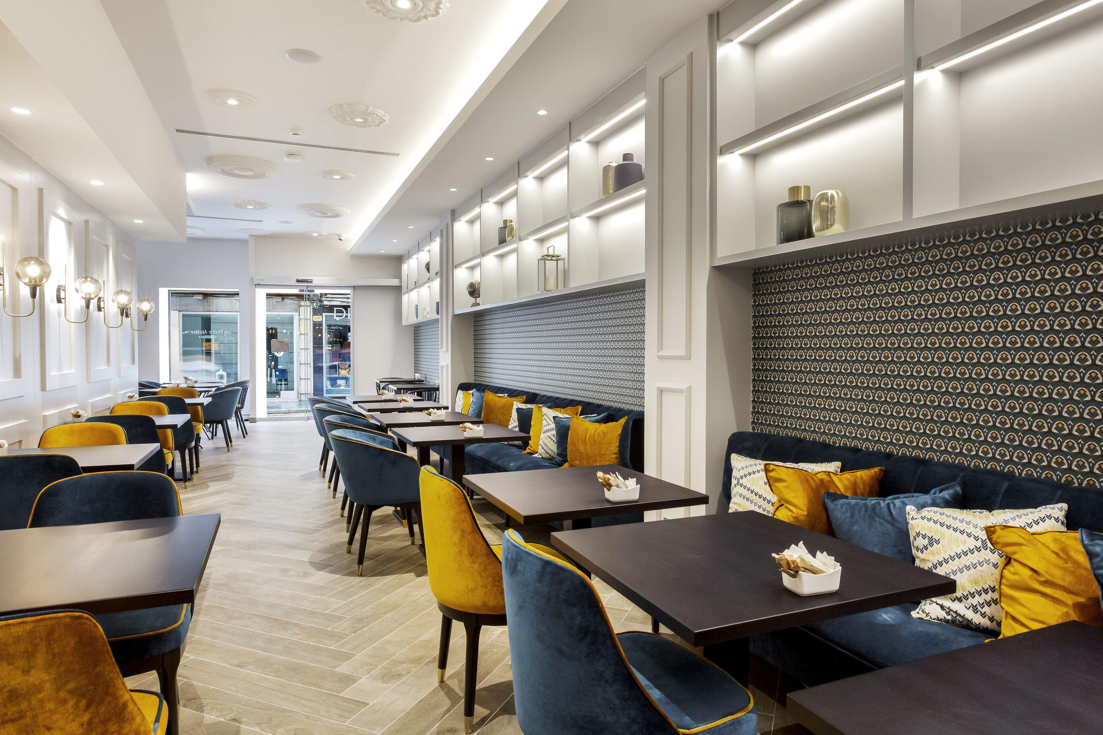 A modern restaurant interior with high tables, blue and yellow upholstered seating, and patterned wall decor.