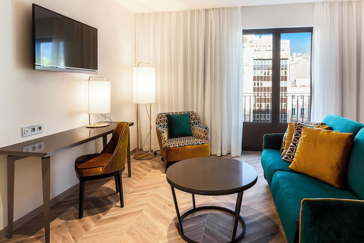 A modern hotel room with a flat-screen TV, a dining table, two chairs, a sofa, and a large window with a city view.