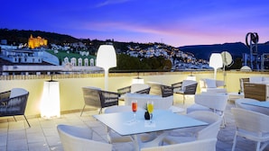 An outdoor terrace with white wicker furniture, a table with drinks, and a view of a town and mountains in the background. - Eurostars Puerta Real Hotel (Granada)