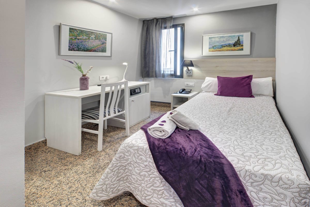 A hotel room with a bed, a desk, a chair, a nightstand, a lamp, and two framed pictures on the wall.