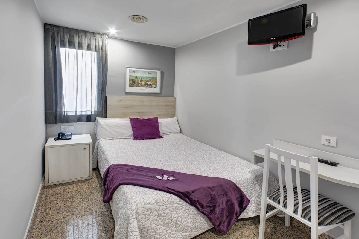 A bedroom with a bed, a bedside table, a desk, a chair, a TV mounted on the wall, and a window with curtains.