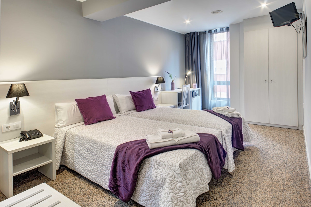 A hotel room with two beds, a desk, a TV, and a window with curtains.