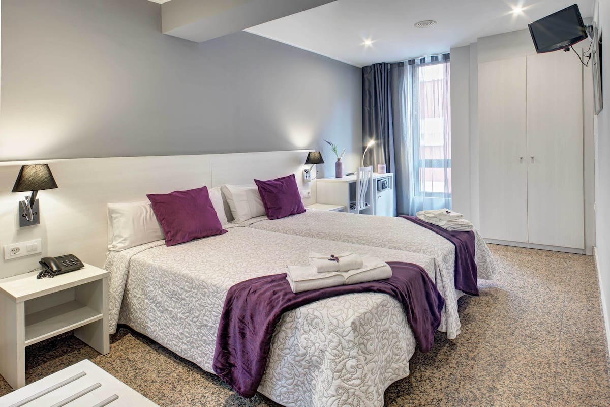 A hotel room with two beds, a desk, a TV, and a window with curtains.