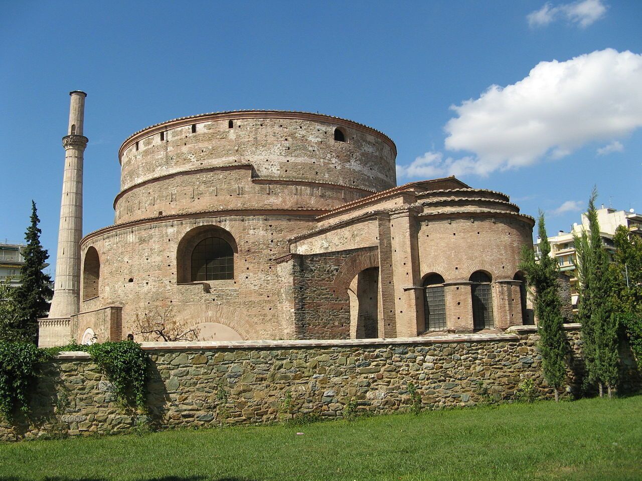 Un bâtiment historique doté d’une tour ronde et de fenêtres en arc, entouré de murs de pierre et de verdure.