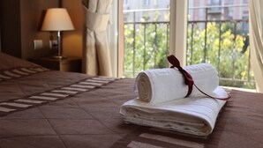 A neatly made bed with a rolled white towel and a red ribbon, a bedside lamp, and a view of a balcony with greenery. - Aegeon Hotel (Thessaloniki)