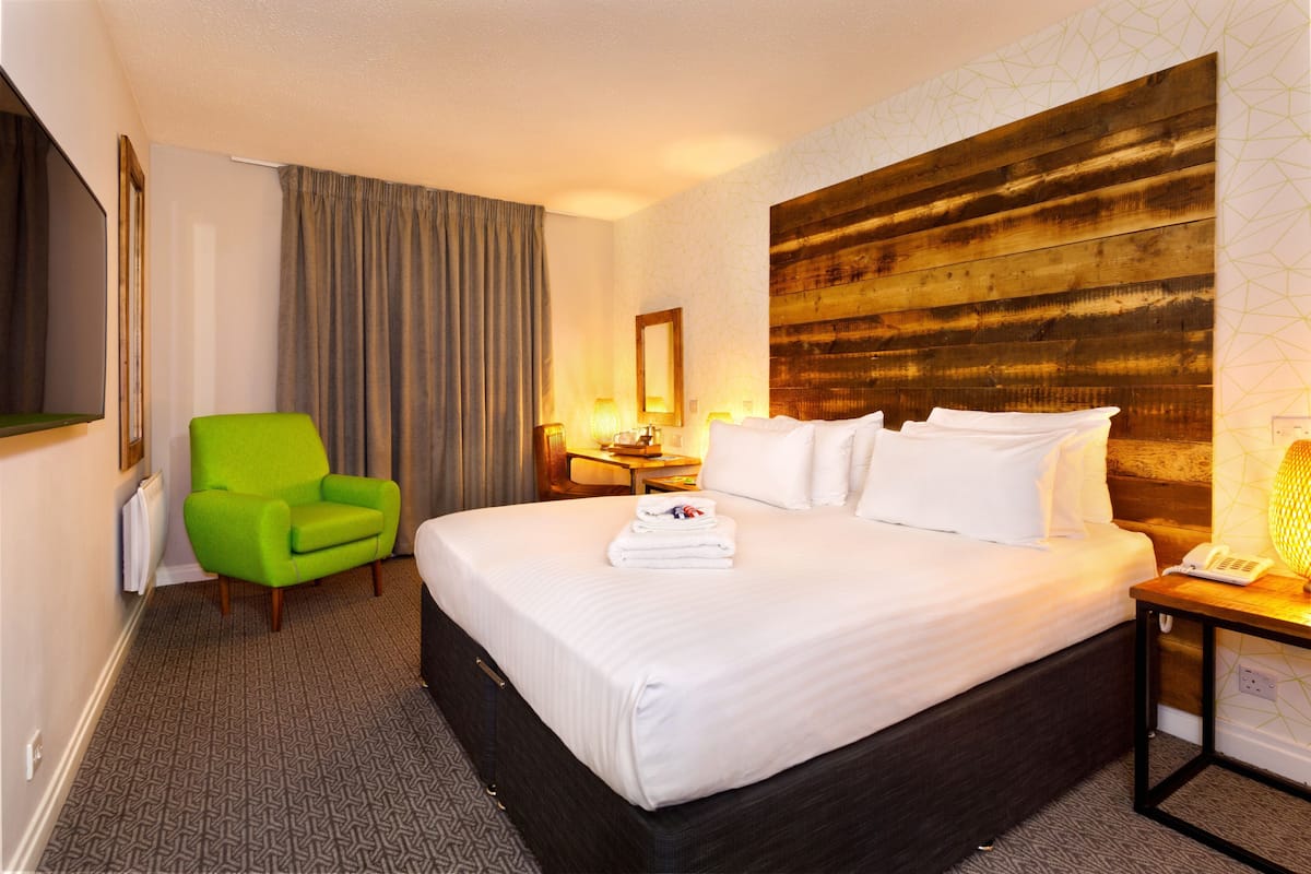 Een hotelkamer met een groot bed, een groene fauteuil, een nachtkastje met een lamp en een houten hoofdeinde.
