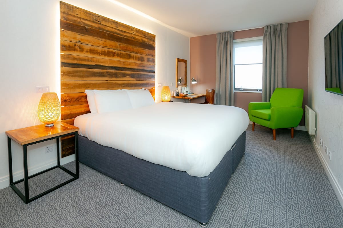 Een hotelkamer met een groot bed, een houten hoofdbord, een bureau met een lamp, een groene stoel en een raam met gordijnen.