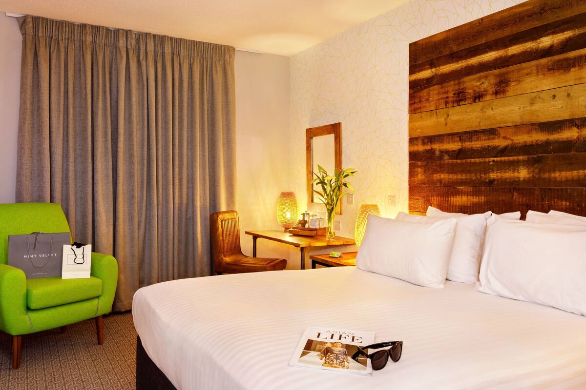 Hotelkamer met een bed, een groene fauteuil, een houten hoofdeinde, een bureau met een lamp en een spiegel.