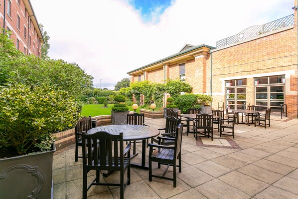 An outdoor patio area with multiple tables and chairs, surrounded by brick buildings and greenery.