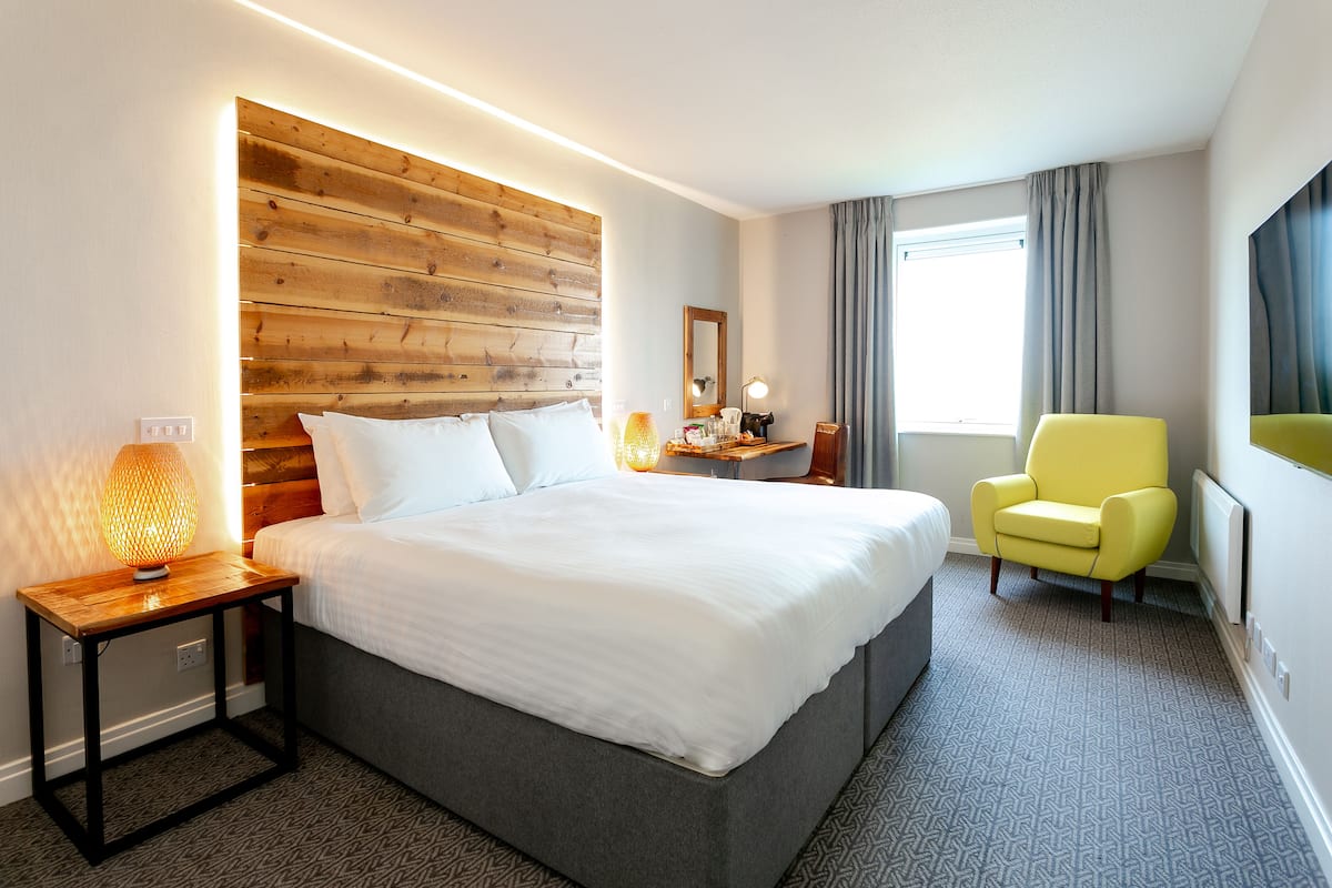 Een hotelkamer met een groot bed, een houten hoofdbord, een nachtkastje met een lamp, een stoel en een raam met gordijnen.
