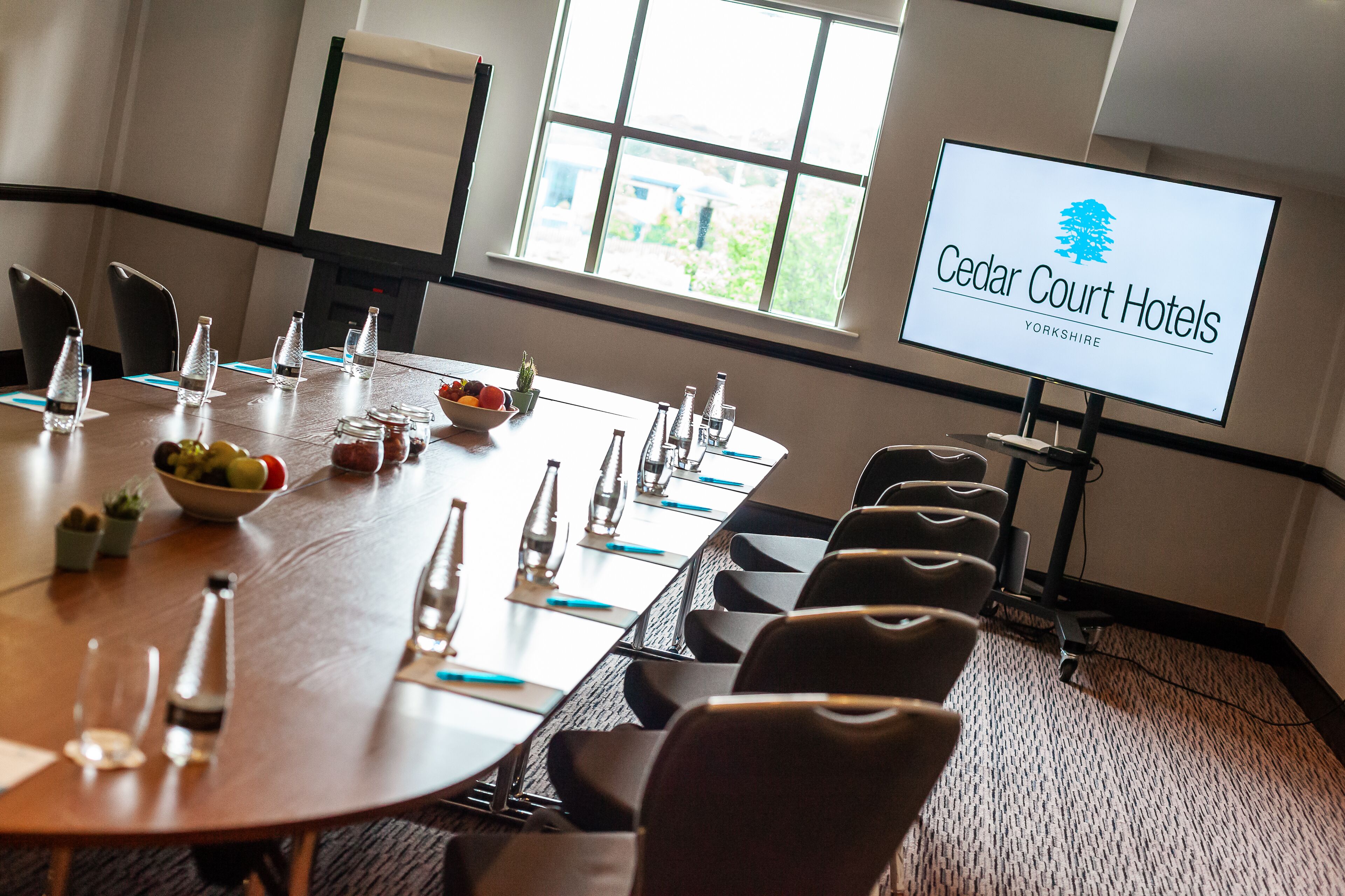 Une salle de conférence de l’hôtel Cedar Court, équipée d’une longue table, de chaises, de bouteilles d’eau et d’un écran affichant le nom de l’hôtel.